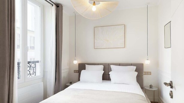 A hotel room with a large bed, a window with curtains, a ceiling fan, a wall-mounted light, and a framed picture. - A Romantic 1-BR with Services in Batignolles (Paris)