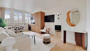Un soggiorno moderno con un divano, un tavolo da pranzo e sedie. È presente un angolo cottura con mobili in legno e un televisore montato a parete.
