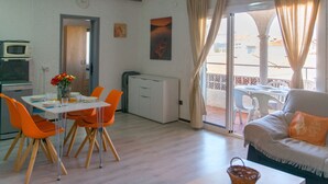 A modern living room with a sofa, a dining table with orange chairs, a kitchen area with a microwave, and a balcony with a table and chairs. - WELCS APARTMENT 367 EMP with communal pool (Empuriabrava)