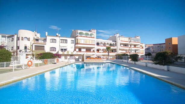 A swimming pool with a clear blue water surface, surrounded by a white building with balconies and a red car parked nearby. - WELCS APARTMENT 367 EMP with communal pool (Empuriabrava)