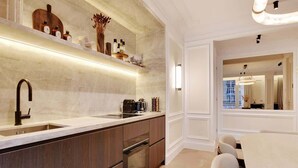 A modern kitchen with dark wood cabinets, a stainless steel sink, and a built-in oven. There is a dining area with a white table and chairs, and a large mirror reflecting the kitchen. - The Lander - 3BR/3BA - Rennes II (Paris)