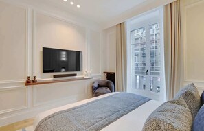 Moderni hotellihuone, jossa on suuri sänky, seinälle asennettu taulutelevisio, ikkuna, josta on näkymä rakennuksiin, ja pieni nojatuoli.