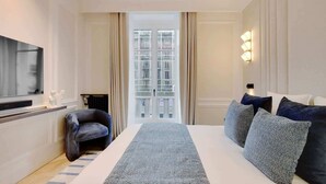 A hotel room with a large bed, a flat-screen TV, a seating area with a chair, and a view of the city through a glass door. - The Lander - 3BR/3BA - Rennes II (Paris)