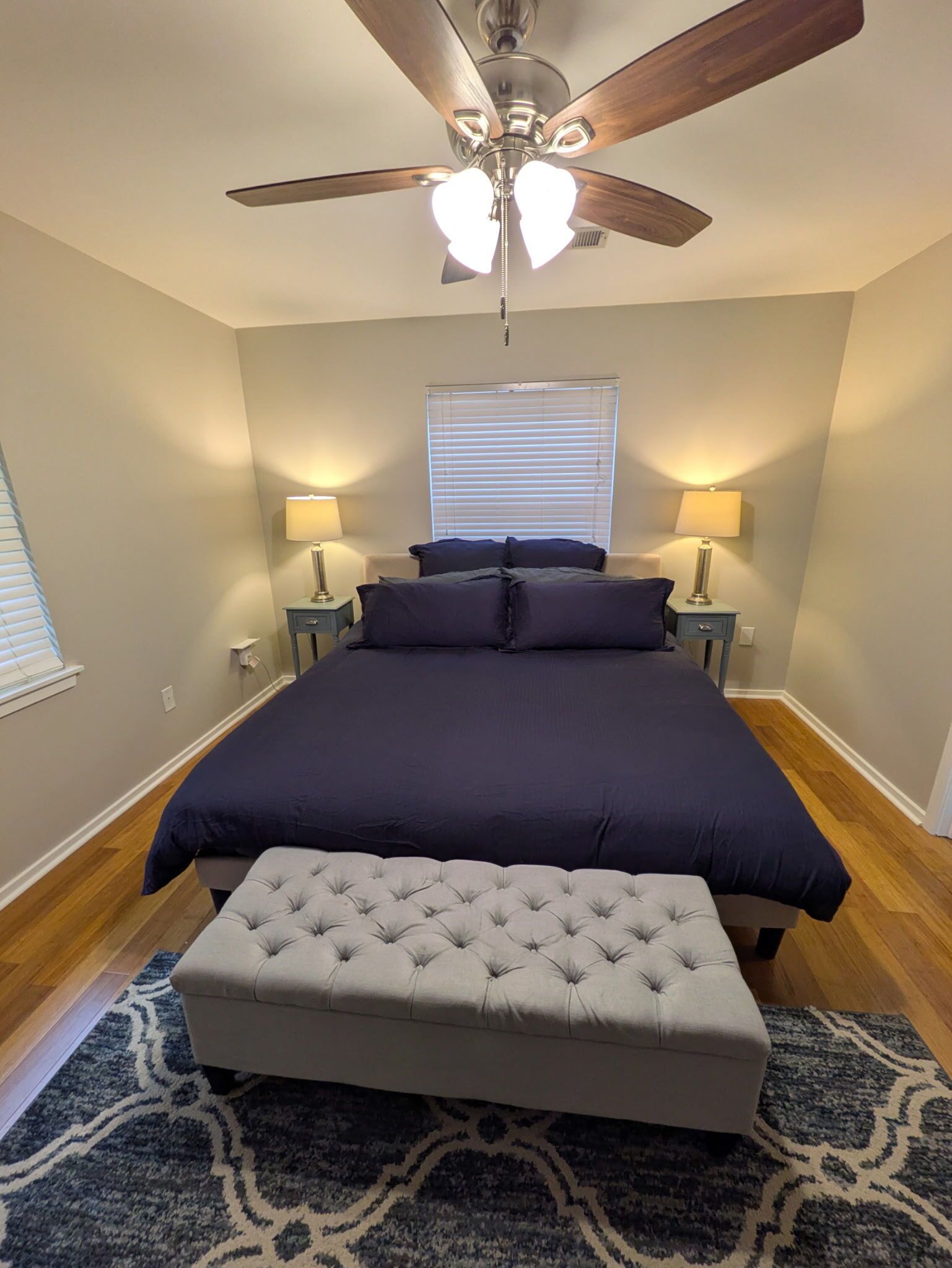 A bedroom with a large bed, two bedside lamps, a ceiling fan, and a window with blinds.