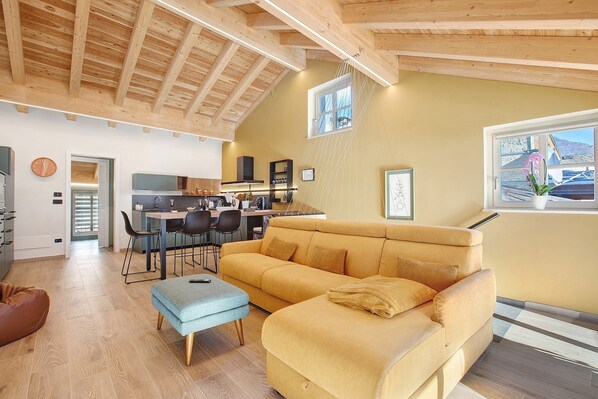 A spacious living area with a sofa, ottoman, and dining table. A kitchen is visible in the background. - La Maison De Ida - Grande Apartment with Mountain View, Wi-Fi and Air Conditioning (Aosta)