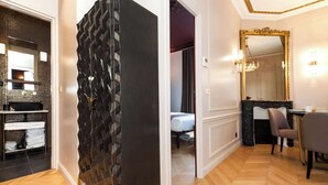 Ein Hotelzimmer mit modernem Design, das eine dunkel strukturierte Trennwand, einen Holzboden, einen Essbereich mit Tisch und Stühlen sowie einen großen Spiegel mit goldener Rahmung aufweist.