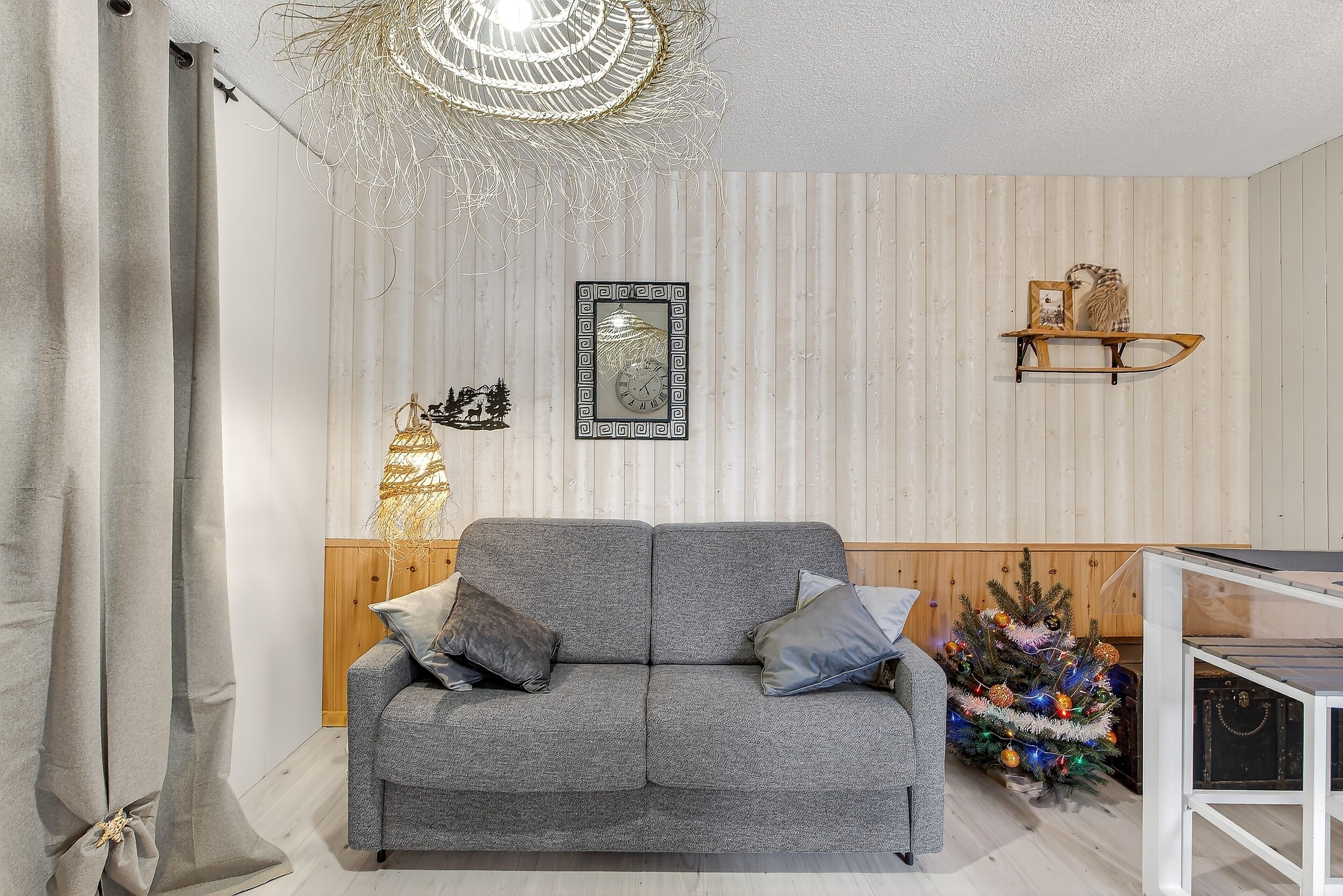 Ein graues Sofa mit Kissen, eine Wanduhr, eine Wanddekoration und ein kleiner Weihnachtsbaum.