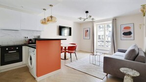 Une cuisine moderne équipée d’un lave-linge, un coin repas avec des chaises rouges et un salon avec un canapé gris.