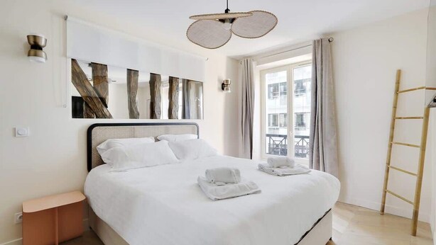 Un lit bien fait, avec du linge de lit et des serviettes blanches, une tête de lit moderne et une grande fenêtre avec des rideaux.