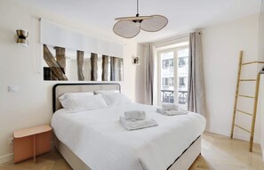 Een netjes opgemaakt bed met witte lakens en handdoeken, een modern hoofdbord en een groot raam met gordijnen.