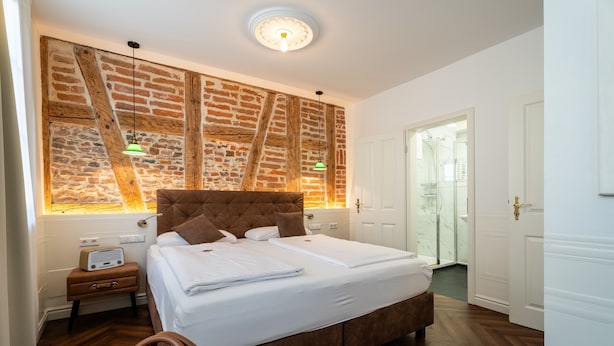 A bedroom with a large bed, a brick wall, a wooden floor, a nightstand with a radio, and a door leading to a bathroom. - "Suite Gentleman" with King-Size Bed and Air Conditioning (Gengenbach)