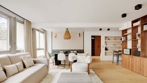 A modern living room with a sofa, chairs, a coffee table, and a dining area with a table and chairs. There is a bookshelf and a TV in the background. - A Refined 3-BR/3BA in Trocadéro - Tour Eiffel (Paris)