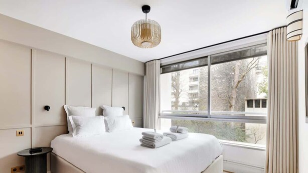 A modern bedroom with a large bed, a bedside table, a lamp, and a window with curtains. - A Refined 3-BR/3BA in Trocadéro - Tour Eiffel (Paris)