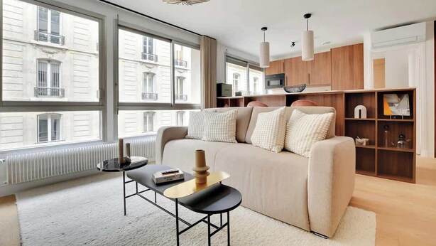 En moderne stue med sofa, sofabord og fjernsyn. Store vinduer giver naturligt lys og udsigt til bygningen udenfor.