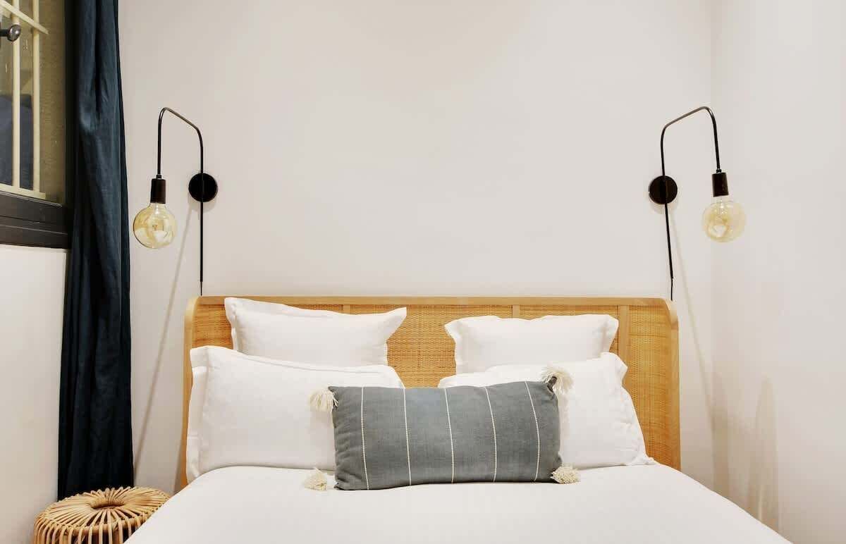 A neatly made bed with white and gray pillows, a wooden headboard, and two wall-mounted light fixtures with exposed bulbs.