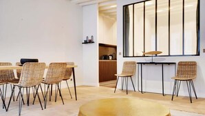 Un espace repas moderne avec des chaises en osier et une table ronde, une grande fenêtre avec des stores verticaux, et une cuisine visible au fond.