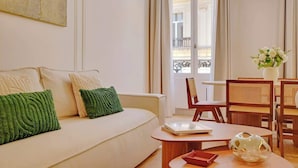 Een moderne woonkamer met een witte bank, groene kussens, een houten salontafel en een eethoek met rieten stoelen en een witte vaas.