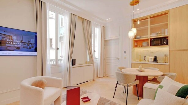 A modern living room with a sofa, a coffee table, and a dining area with a table and chairs. There is a kitchen area with a microwave and a shelf with dishes. - An Elegant 1-BR/1Ba with Services in La Madeleine (Paris)