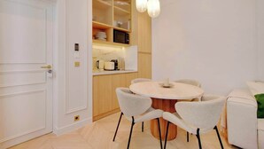 A modern kitchen with white cabinetry, a microwave, and a coffee maker on the countertop. There is a round dining table with four chairs and a hanging light fixture above. - An Elegant 1-BR/1Ba with Services in La Madeleine (Paris)