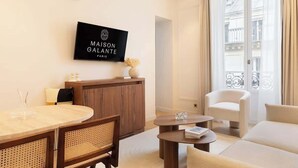 A modern hotel room with a wooden reception desk, a flat-screen TV displaying the hotel name, a round dining table with chairs, a sofa, and a small coffee table. - A Sophisticated 1-BR with Services in La Madeleine (Paris)