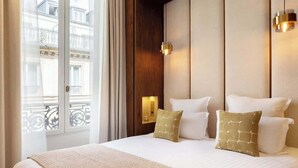 A hotel room with a large bed, a wooden headboard, two wall-mounted lights, and a view of buildings through a window. - A Sophisticated 1-BR with Services in La Madeleine (Paris)