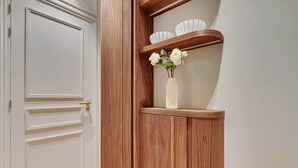 Eine weiße Tür mit goldener Klinke, ein Holzregal mit weißen Schüsseln und einem Blumenstrauß, sowie eine weiße Wand.
