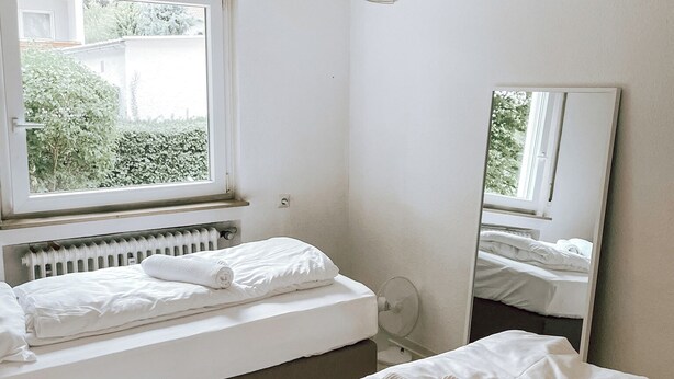 Ein Schlafzimmer mit Bett, Fenster, Heizkörper, Spiegel und einer Deckenleuchte.