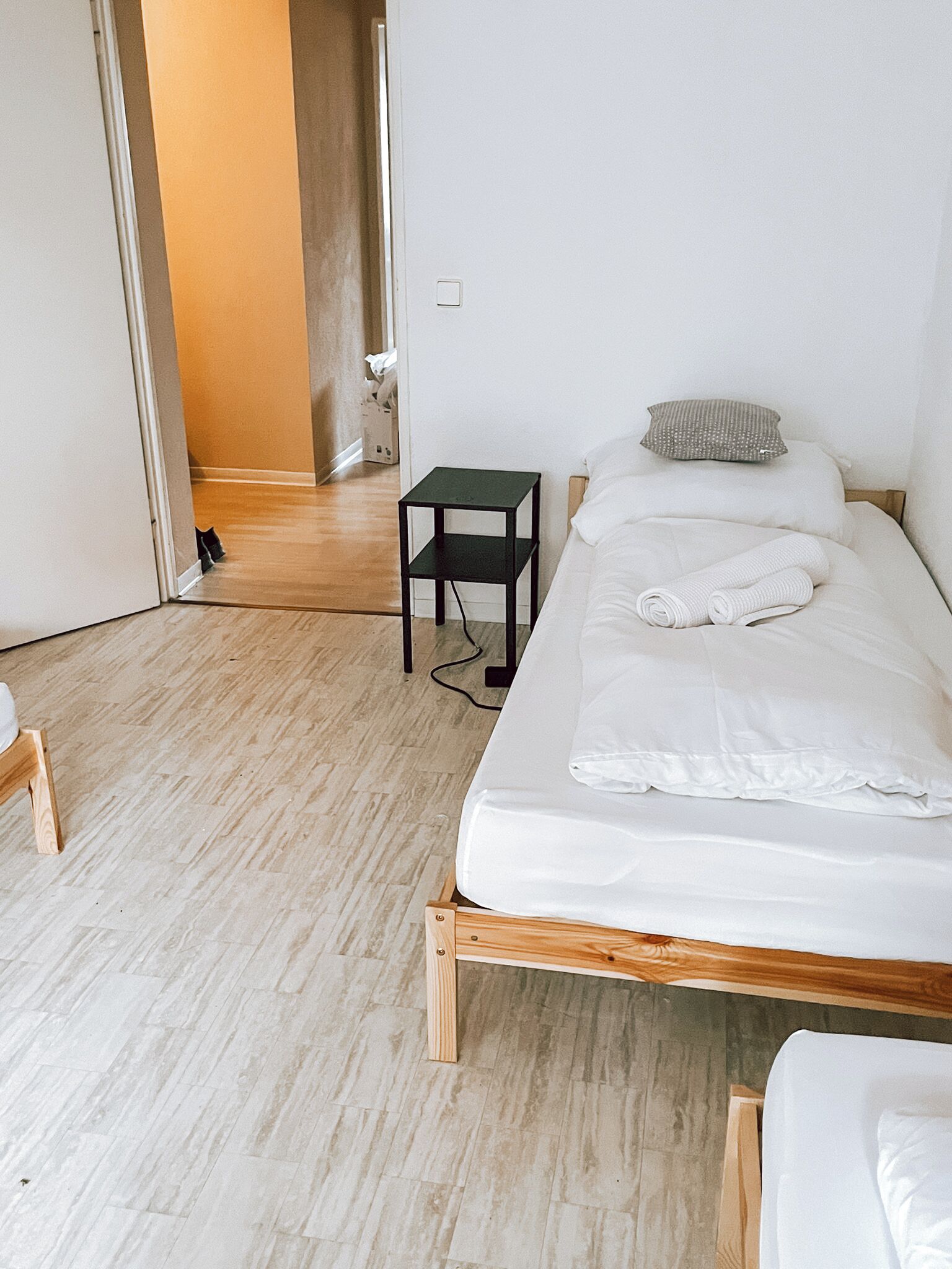 A bedroom with a wooden bed frame, white bedding, a small black nightstand, and a wooden chair.