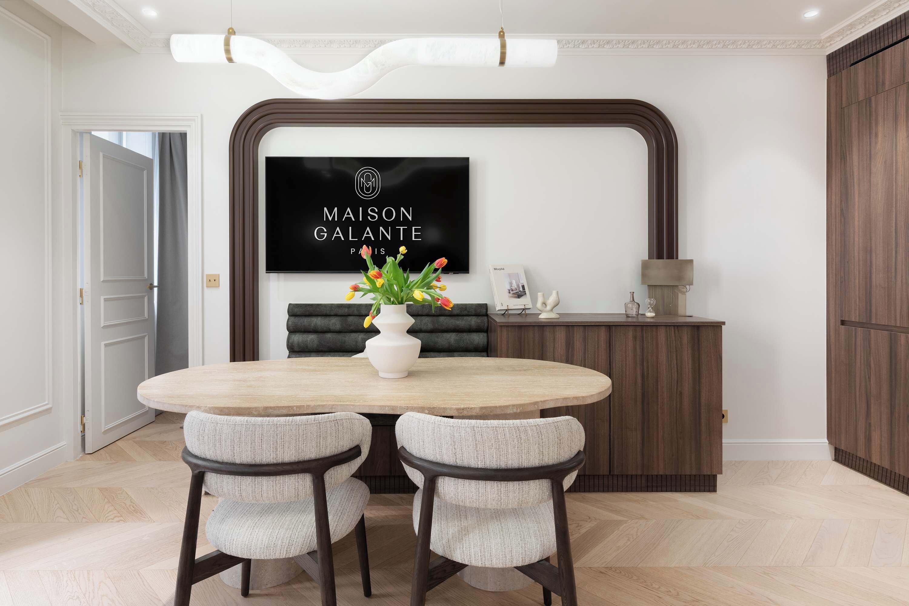 A dining area with a round table, two chairs, a vase with flowers, a dark upholstered bench, a wooden cabinet, and a framed sign on the wall.