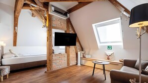 A loft-style room with exposed wooden beams, a brick wall, a bed, a TV, a lamp, and a window. - "Suite Heimat" with King-Size Bed and Air Conditioning (Gengenbach)