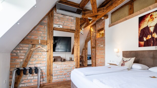 A bedroom with a sloped ceiling, exposed brick walls, a wooden ceiling, a bed, a hanging coat rack, and a painting on the wall. - "Suite Heimat" with King-Size Bed and Air Conditioning (Gengenbach)