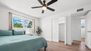 Un dormitorio con cama, ventilador de techo, ventana con cortinas y vistas al exterior.