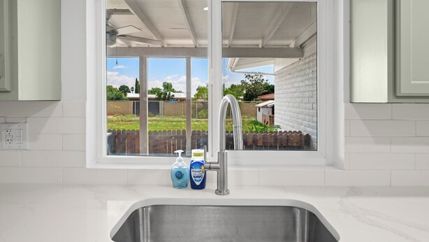 Cocina con ventana, pileta de acero inoxidable y vista a una casa de ladrillo y vegetación.