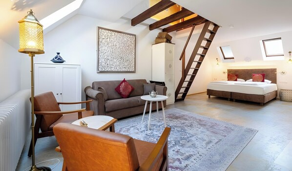 Une pièce de style « loft », avec un plafond haut, des poutres apparentes, un lit en mezzanine, un canapé, une table basse et un lampadaire.