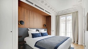 A modern bedroom with a large bed, wooden headboard, and a view of the city through large windows. - ONIRI - Serviced 1-BR - Place Vendôme Louvre III (Paris)
