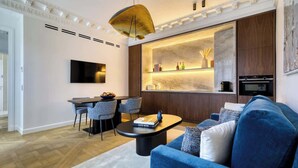 A modern living room with a blue sofa, a dining area with a table and chairs, a kitchen area with a built-in oven and microwave, and a large TV mounted on the wall. - ONIRI - Serviced 1-BR - Place Vendôme Louvre III (Paris)