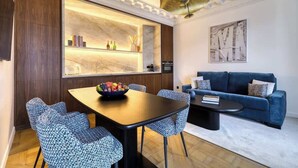 A modern living room with a dark blue sofa, a round coffee table, and a dining area with a wooden table and patterned chairs. - ONIRI - Serviced 1-BR - Place Vendôme Louvre III (Paris)