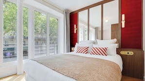 A hotel room with a large bed, a red accent wall, and a view of buildings through large windows. - ONIRI - Somptuous Serviced 3-BR - Saint-Germain (Paris)