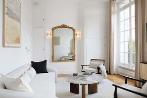 Uma sala de estar espaçosa com um sofá branco, uma mesa de centro redonda e um espelho grande com moldura dourada.