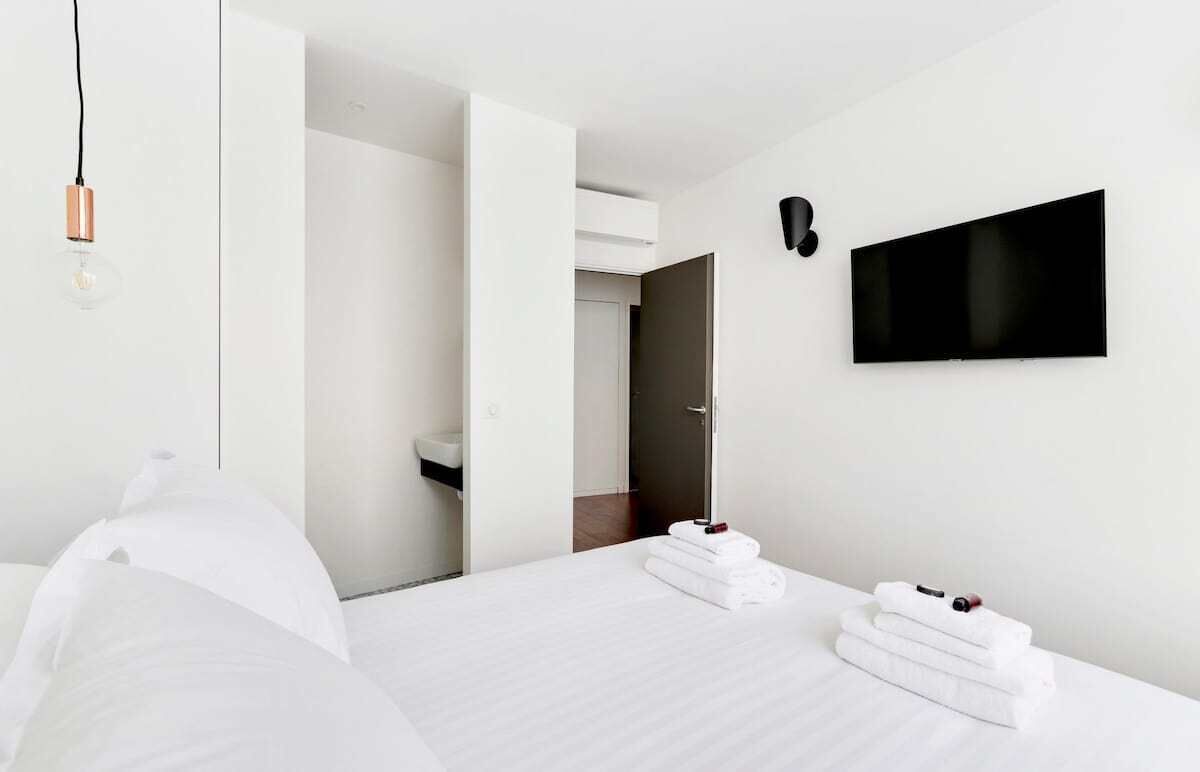 A modern hotel room with a bed, a flat-screen TV mounted on the wall, and a bedside table with towels.