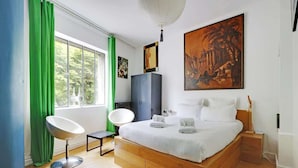 A modern bedroom with a large bed, green curtains, a white chair, a wooden floor, and a painting on the wall.
