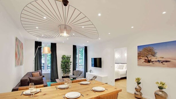 Una sala de estar moderna con comedor, un ventanal grande, un televisor de pantalla plana y un cuadro de un paisaje desértico.