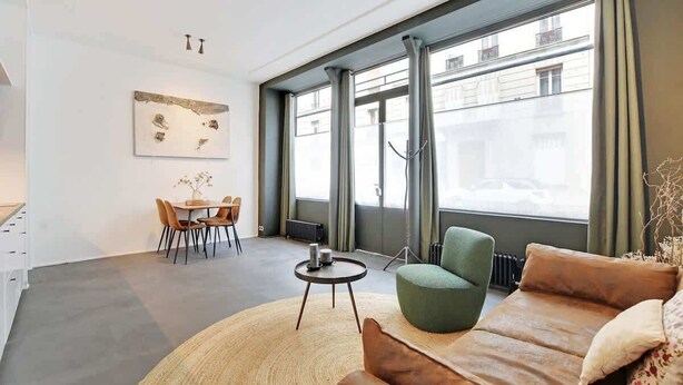 Un salon moderne avec un canapé, une table à manger et des chaises, ainsi qu’une grande fenêtre avec des rideaux.