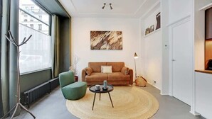 Un salon moderne avec un canapé marron, un fauteuil vert et une table basse ronde. Un grand tableau abstrait orne le mur et un radiateur est posé sur le sol.