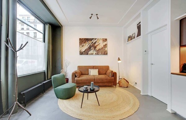 Uma sala de estar moderna com um sofá castanho, uma poltrona verde e uma mesa de centro redonda. Há um quadro abstrato grande na parede e um radiador no chão.