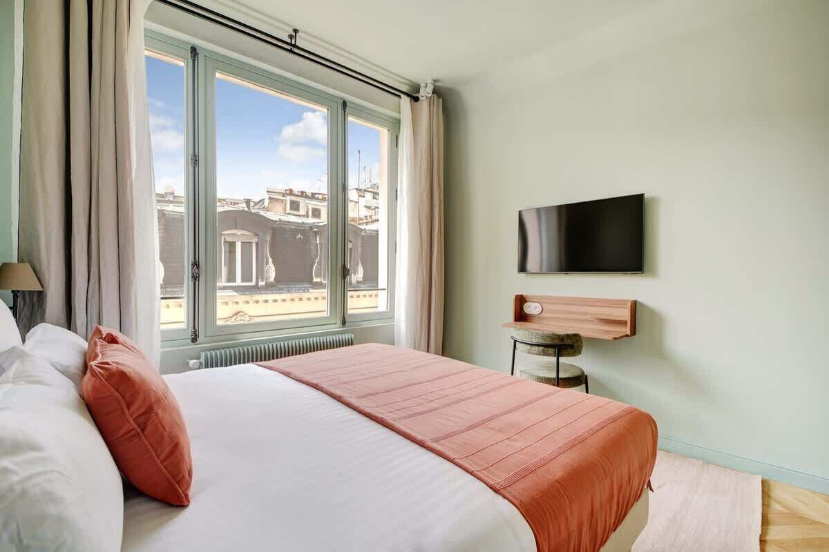 A hotel room with a large bed, a TV mounted on the wall, and a window with curtains.