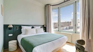 Ein modernes Schlafzimmer mit einem großen Bett, Blick auf die Stadt durch ein Fenster und einer kleinen Sitzecke.