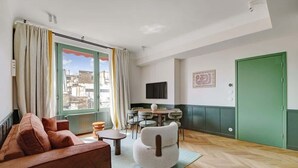 Ein Wohnzimmer mit einer Couch, Sesseln, einem Esstisch und Stühlen. Ein Fenster mit Vorhängen ist sichtbar, und im Hintergrund befindet sich eine grüne Tür.