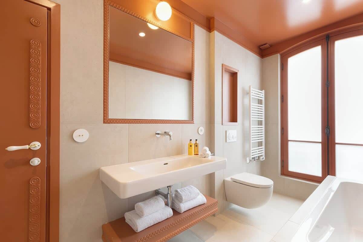 A modern bathroom with a large mirror, white sink, and bathtub. There is a towel rack and a toilet visible.