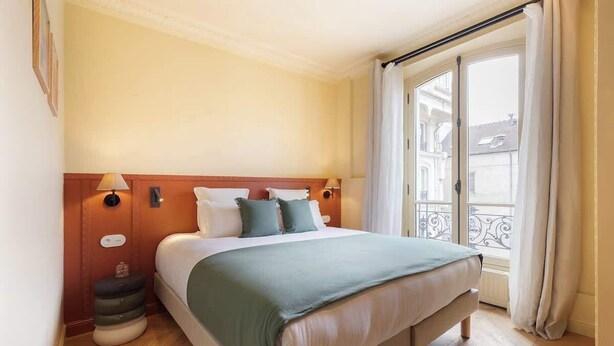 Habitació d'hotel amb un llit gran, una tauleta de nit, una finestra que mostra edificis i un balcó amb barana.